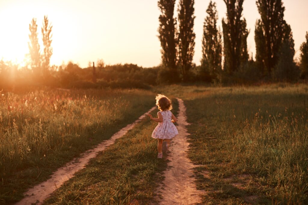 Little cute girl in summer dress runs away into the green blooming fields. Beautiful nature sunset